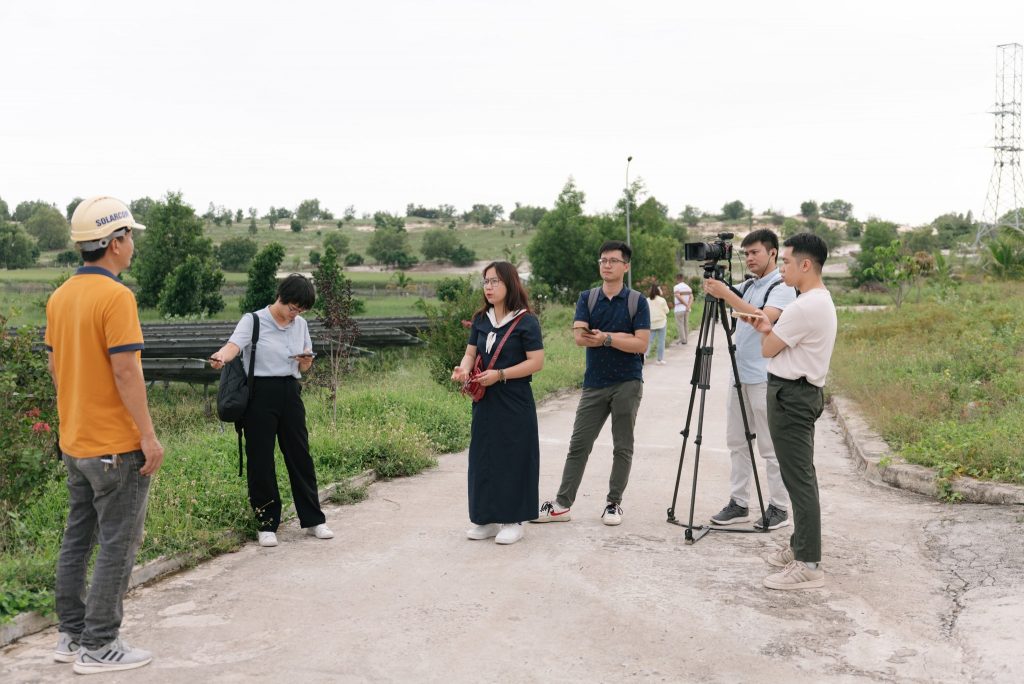 Group of six people on a path, one person wearing a "SOLARCOM" cap, with a camera set up on a tripod. Grass and hills are in the background.