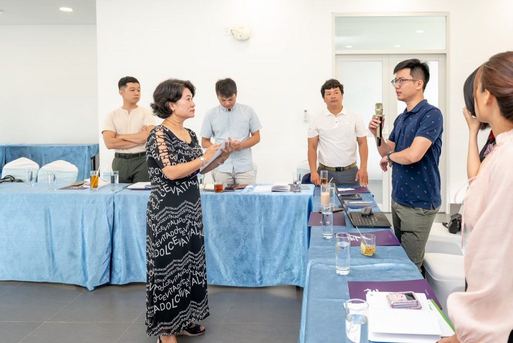 A group of people stand and talk around tables with blue tablecloths in a conference room.